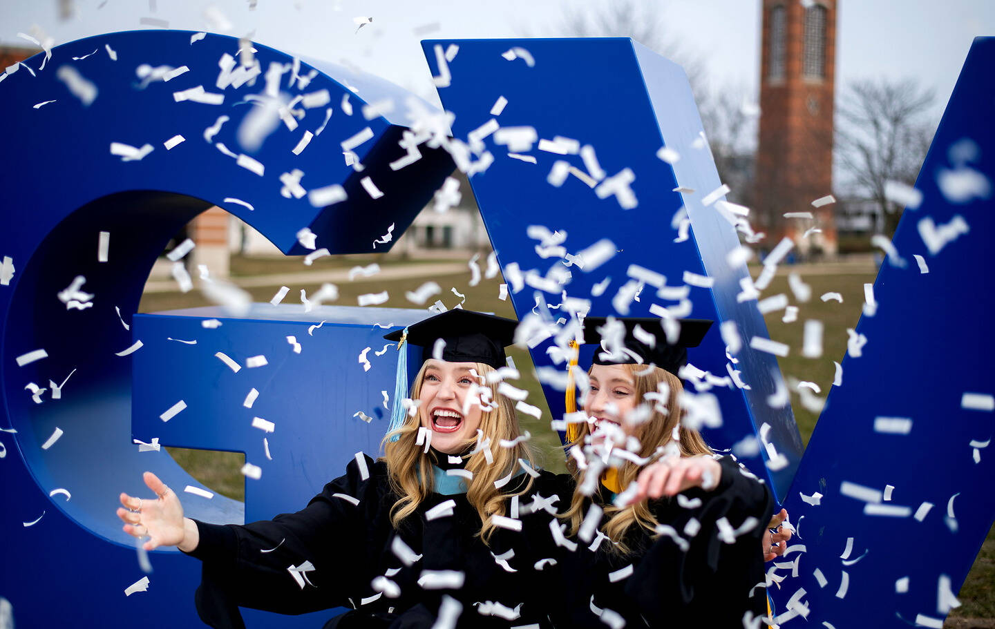 Two graduates in caps and gowns laugh joyfully as white confetti falls around them in front of large blue letters.